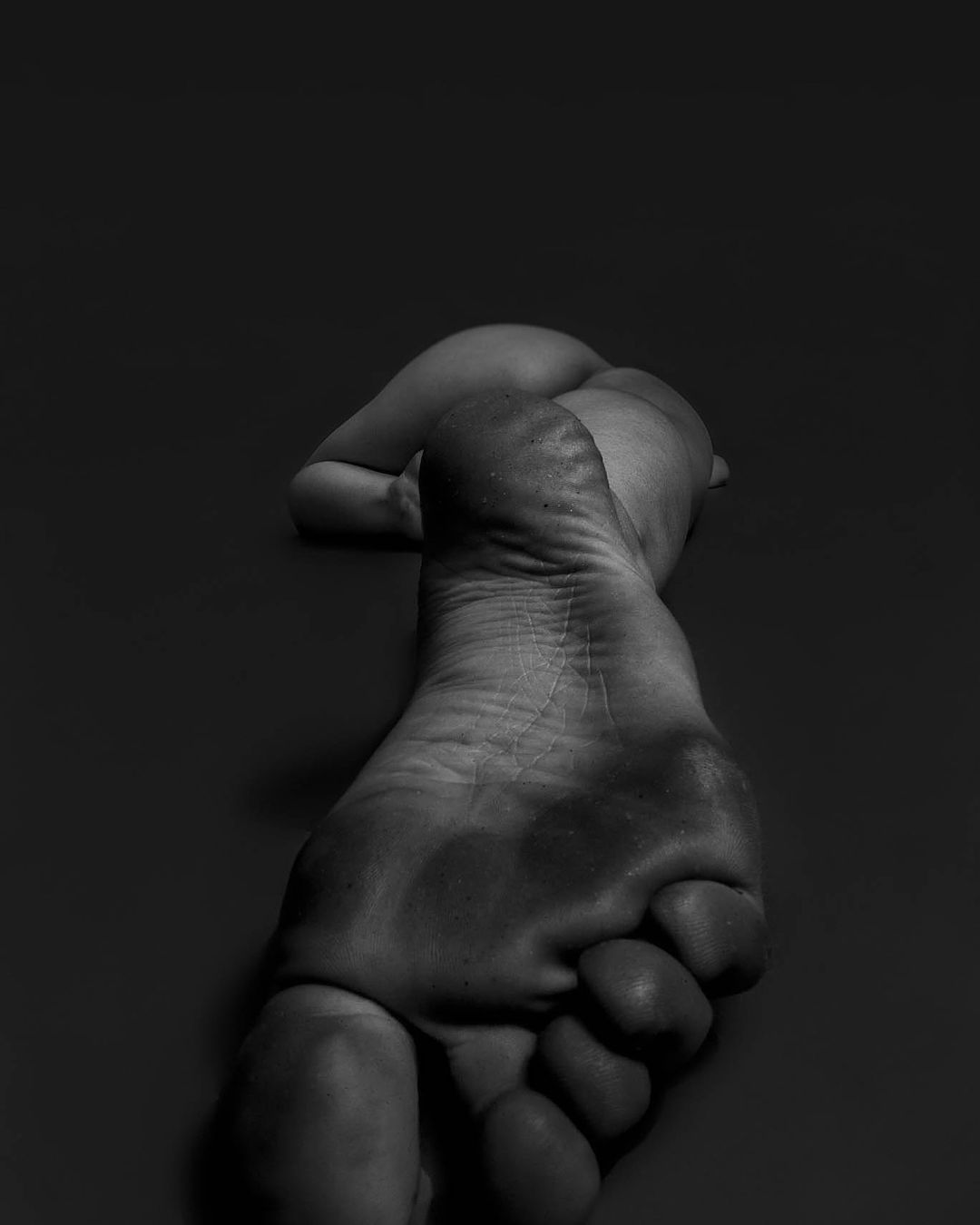Close-up black and white studio shot of a bare foot and partial human figure against a dark background, showcasing textures.