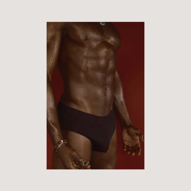 Adult Black man with muscular physique poses shirtless against red backdrop. He is wearing a black brief, a bracelet and necklace.