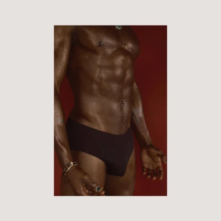 Adult Black man with muscular physique poses shirtless against red backdrop. He is wearing a black brief, a bracelet and necklace.