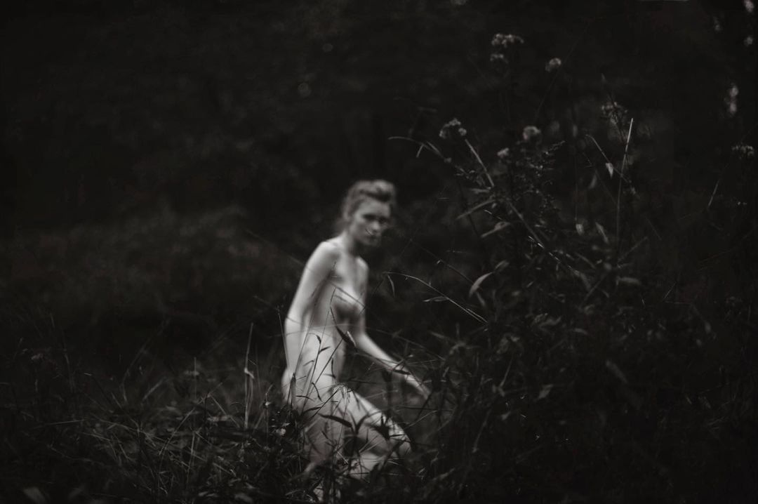 Black and white outdoor portrait of an adult female figure walking through tall grass in an overgrown field with out-of-focus background.
