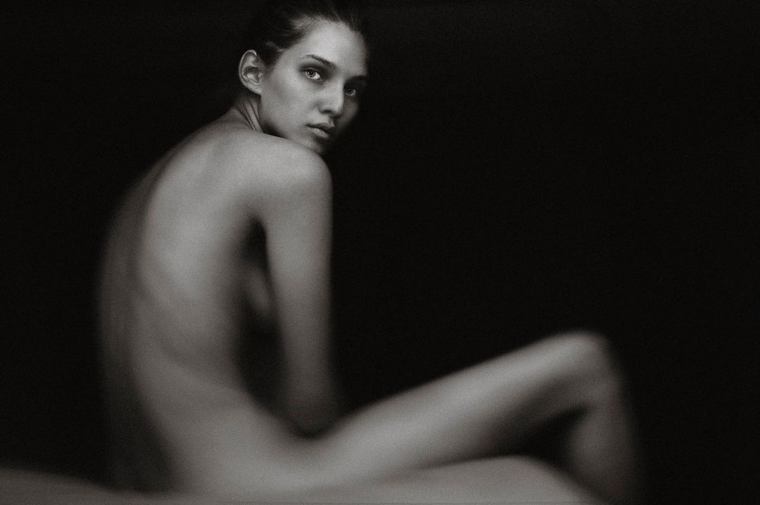 Striking studio portrait of a bare-shouldered young adult female with a slight over-the-shoulder gaze against a dark background.
