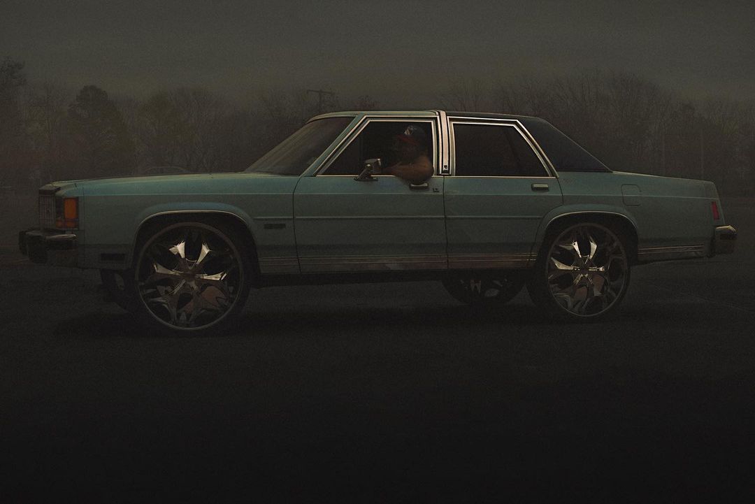 Vintage light blue car with custom chrome wheels parked on a dark street with trees in the distance in a moody outdoor setting.