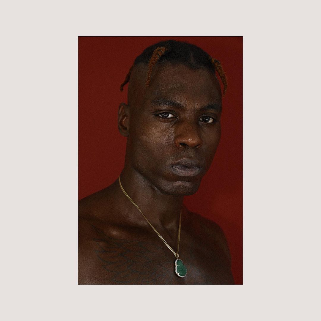 Portrait of a Black man with braided hair wearing a gold chain necklace against a solid red background