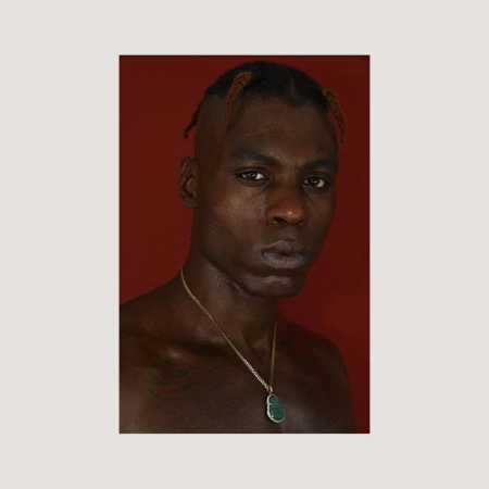 Portrait of a Black man with braided hair wearing a gold chain necklace against a solid red background