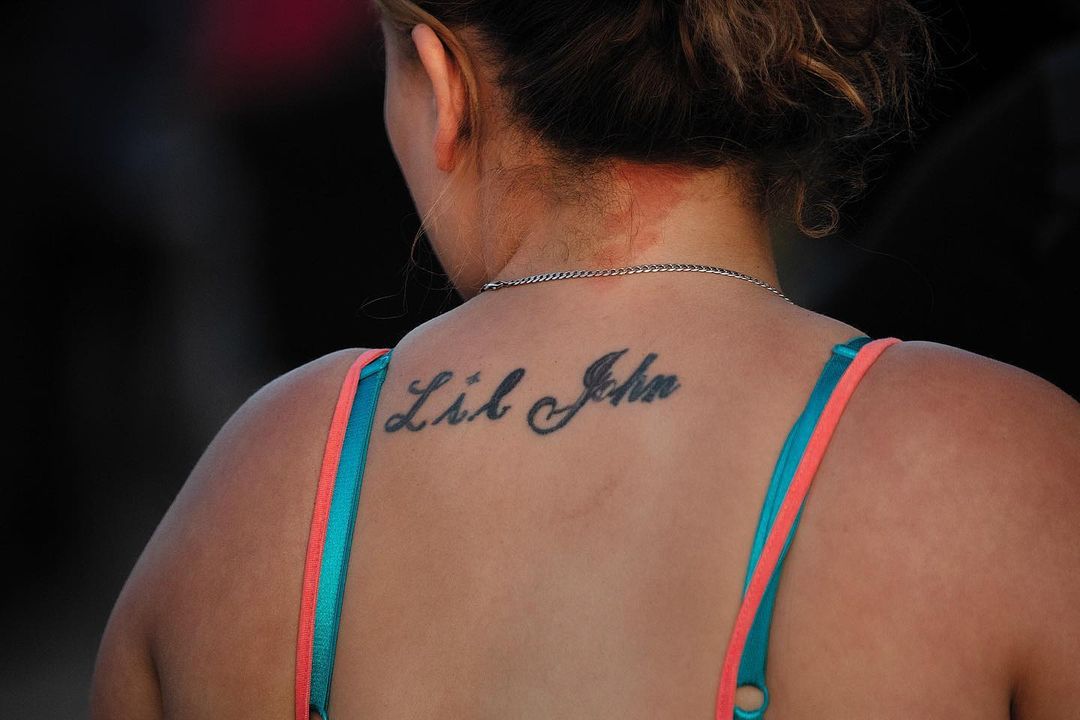 Close-up shot of a woman's back and shoulders featuring a tattoo of the name Lil John, with turquoise and coral colored straps.