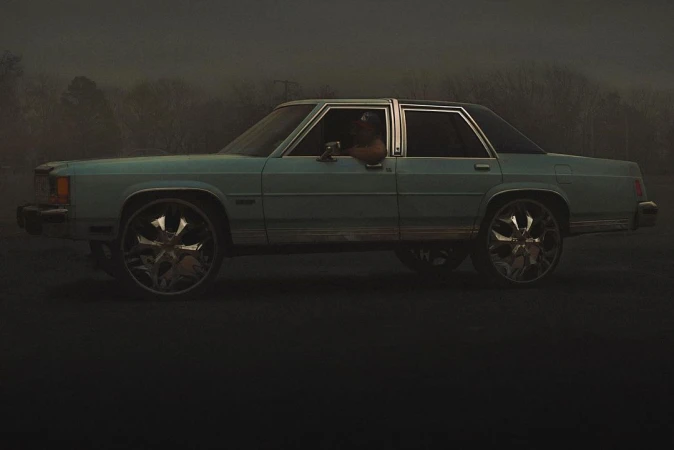 A light blue sedan with large chrome rims parked under an overcast sky with a person visible inside.