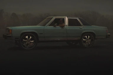 A light blue sedan with large chrome rims parked under an overcast sky with a person visible inside.