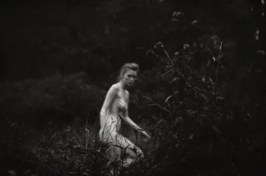 Monochrome portrait of a woman partially obscured by tall wild grasses and foliage outdoors.