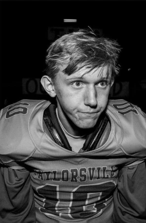 Portrait of a young male American football player wearing a numbered jersey with pads against a dark background.
