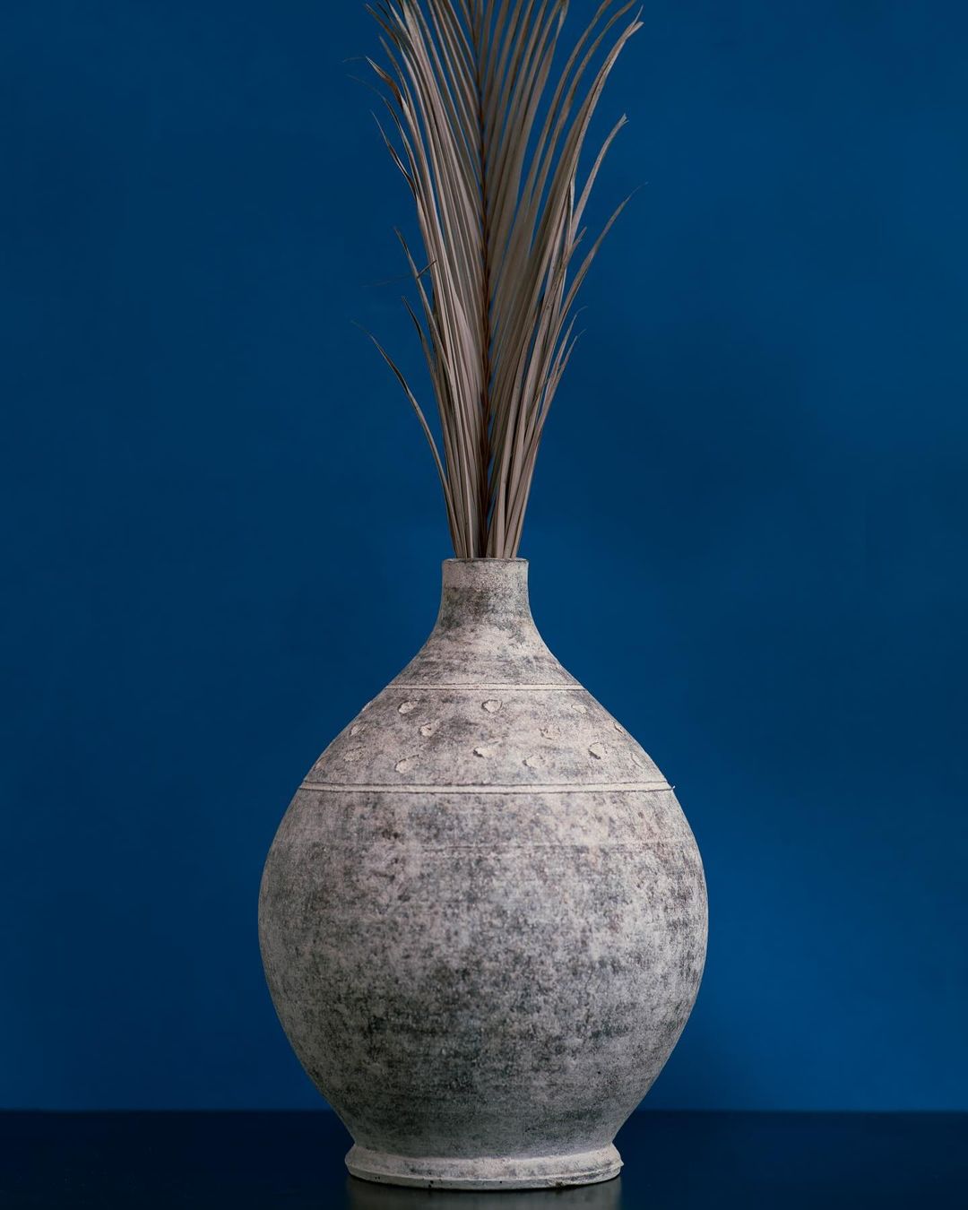 Elegant studio shot of a gray vase with dried floral arrangement against a dark blue background, showcasing simple design and texture.