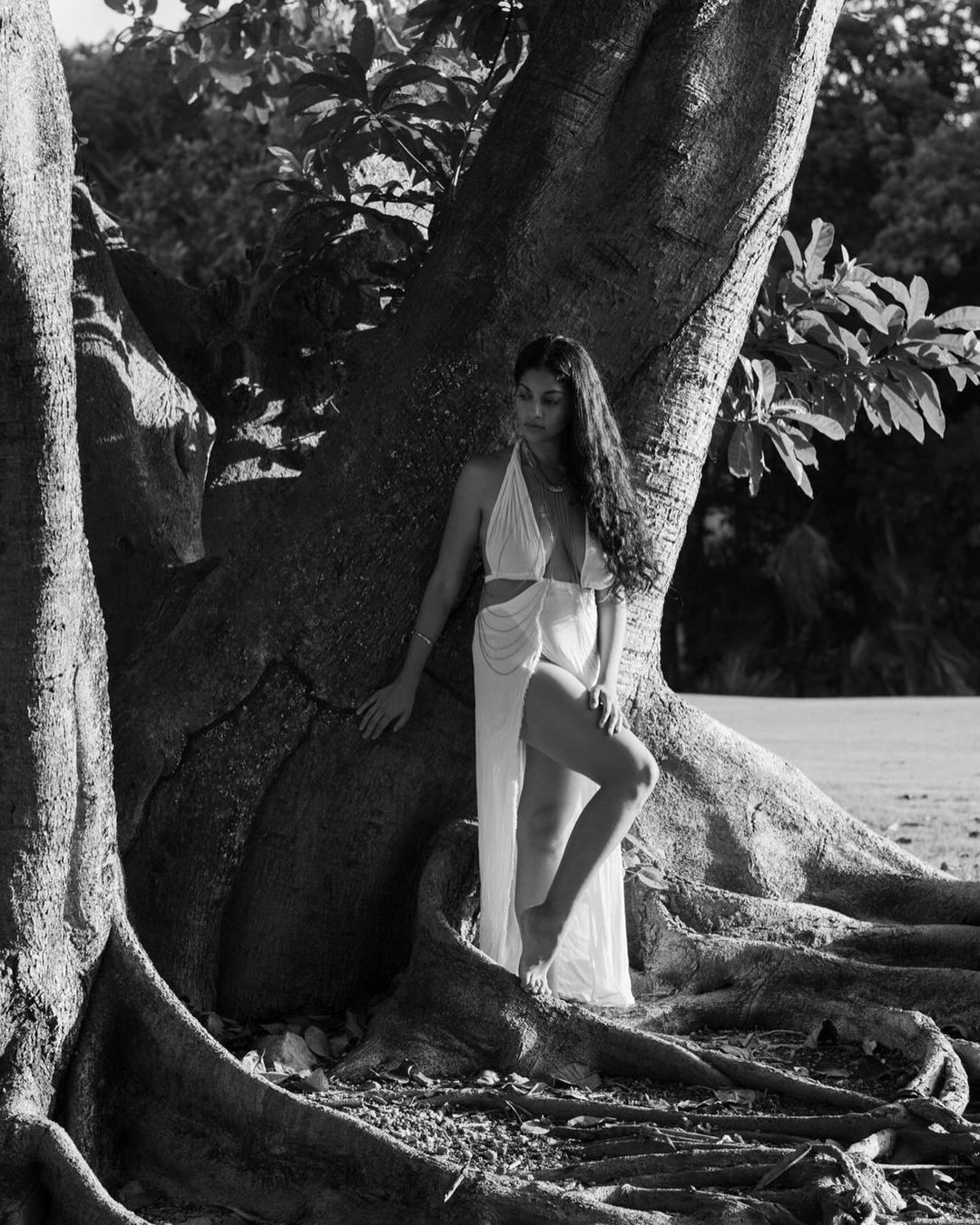 Black and white portrait of a barefoot woman in a white dress, posing against a large tree with roots, on a sandy beach.