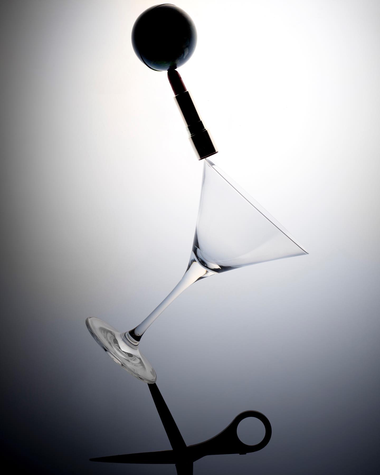 Abstract still life featuring a cocktail glass, lipstick, black sphere, and scissors balanced against a gray gradient background.