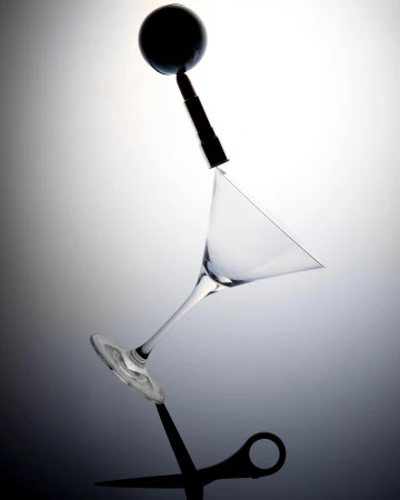 Abstract still life featuring a cocktail glass, lipstick, black sphere, and scissors balanced against a gray gradient background.