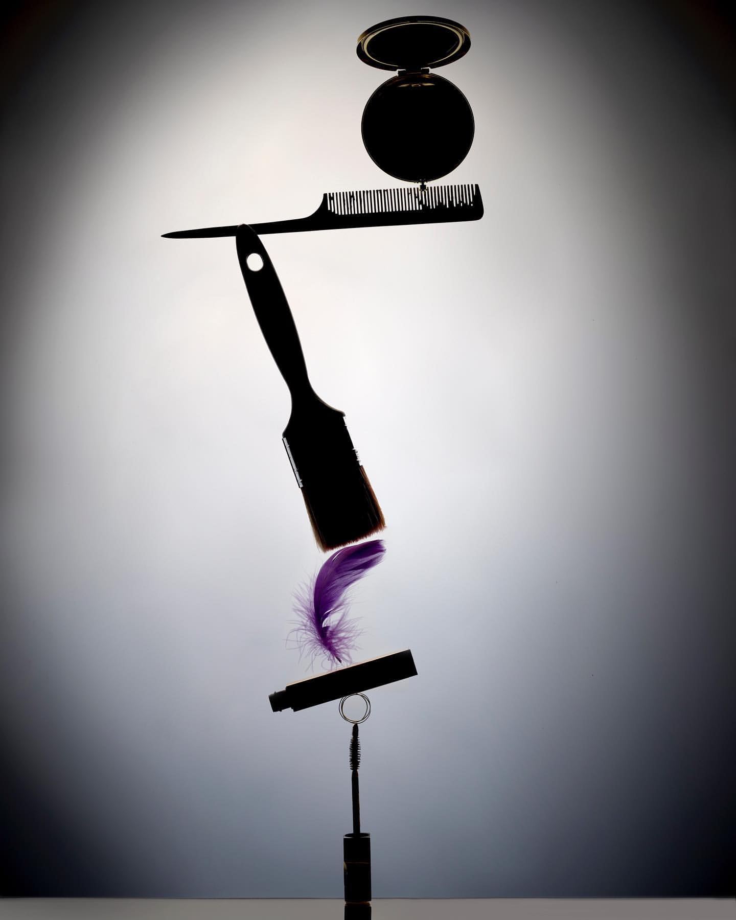 Makeup and hair accessories, including a comb, compact, mascara, and feather, balanced in a vertical stack against a soft gradient background.
