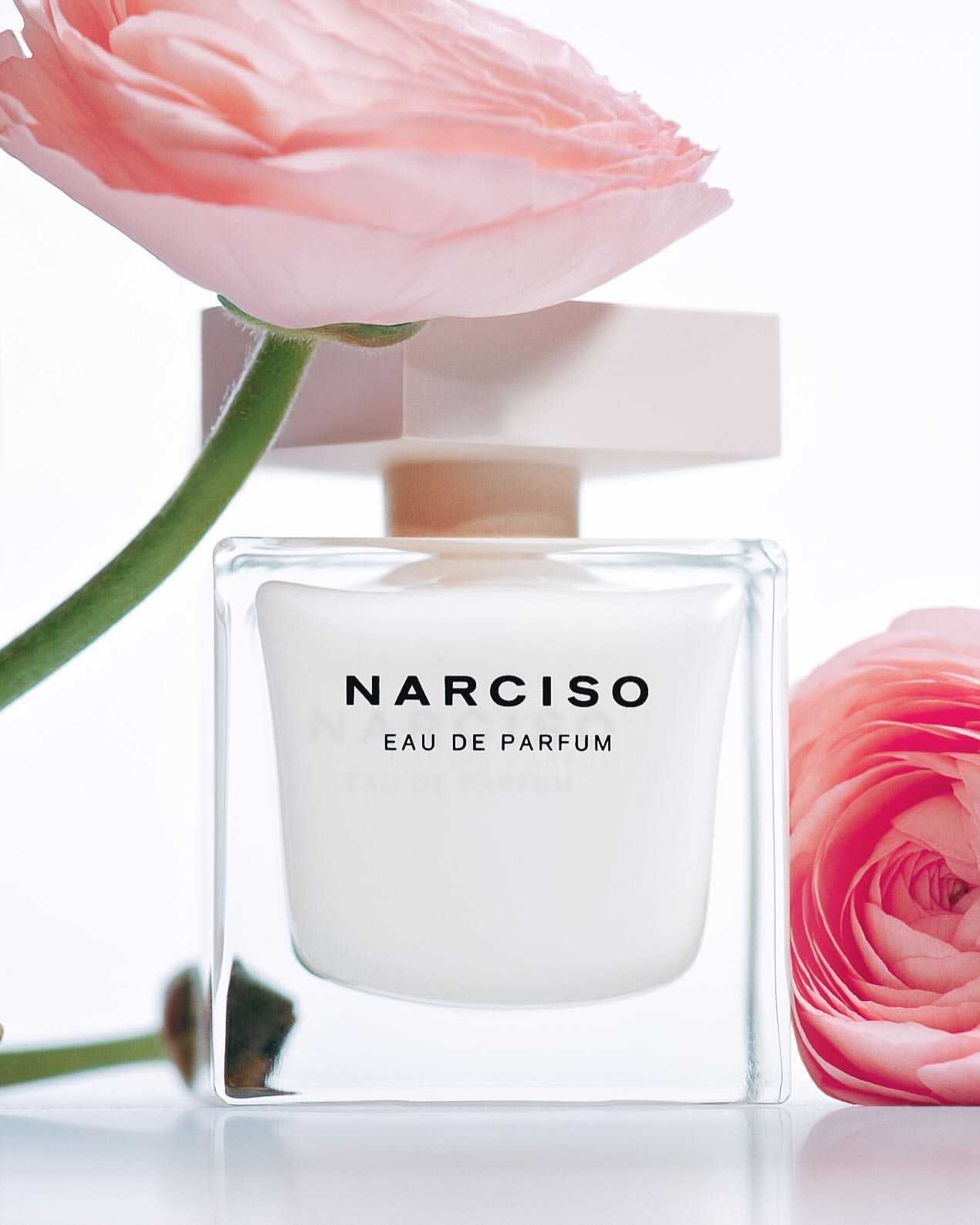 Close-up studio shot of Narciso Eau de Parfum in a clear square bottle with a light pink ranunculus flower in the background.