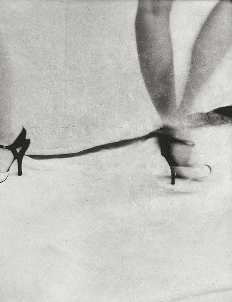 Monochrome studio shot of an adult female model wearing high heels, posing with legs crossed against a light background.