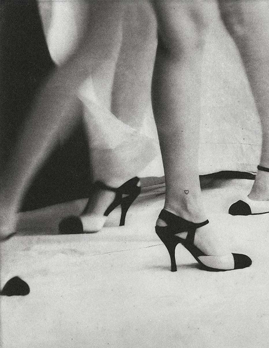 Black and white image of two women's legs wearing black and white high heels, one with a tiny heart tattoo on her ankle.