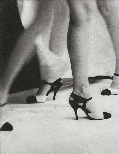 Black and white image of two women's legs wearing black and white high heels, one with a tiny heart tattoo on her ankle.