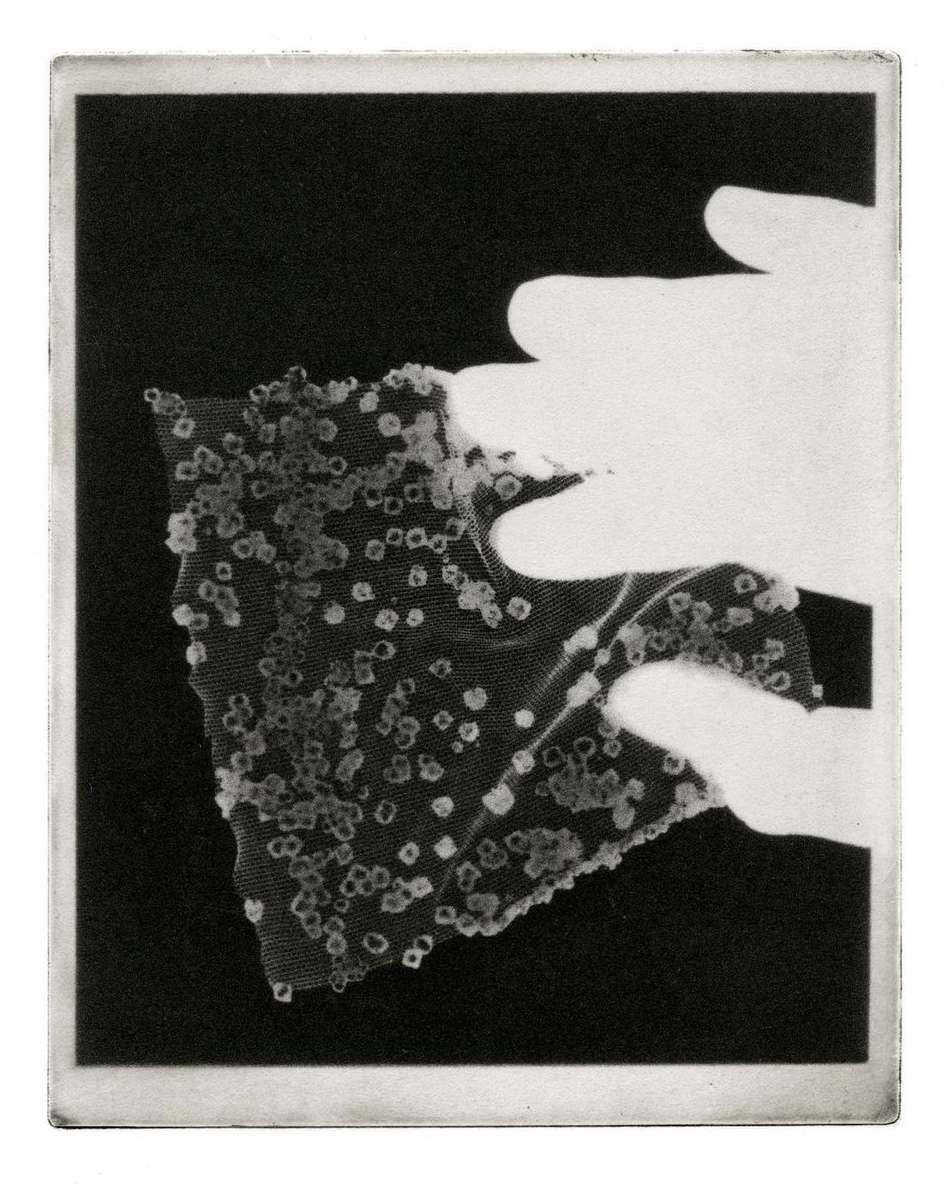 Hands holding a translucent fabric with square embellishments, set against a dark backdrop in a high-contrast, monochrome image.