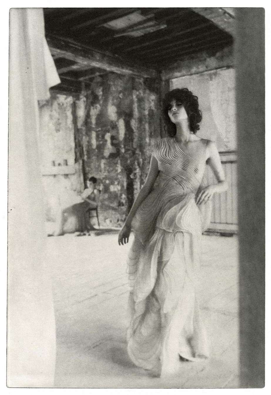 Monochrome fashion shot of a woman posing in a formal layered dress, with another woman sitting in the background.