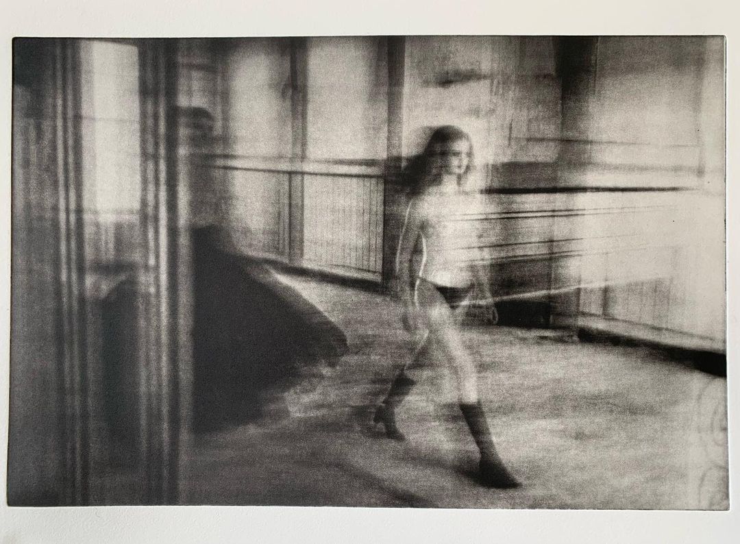 Blurry black and white long exposure photo of a woman walking in an empty interior with textured walls and soft, diffused light.