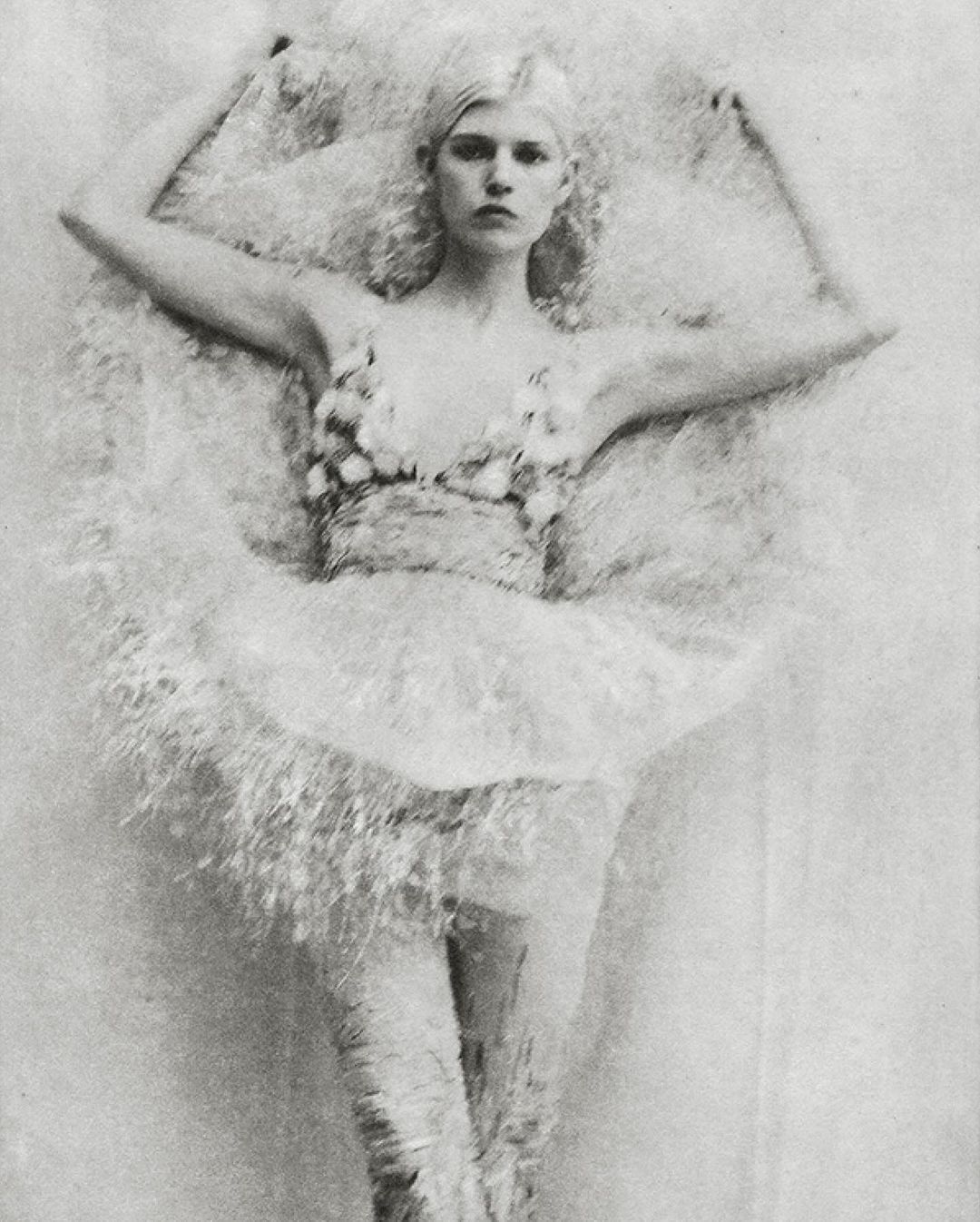 Monochrome portrait of a fair-skinned woman with platinum hair in elaborate, floral-embellished attire, lying against a textured backdrop.