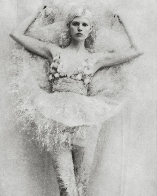 Monochrome portrait of a fair-skinned woman with platinum hair in elaborate, floral-embellished attire, lying against a textured backdrop.
