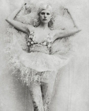 Monochrome portrait of a fair-skinned woman with platinum hair in elaborate, floral-embellished attire, lying against a textured backdrop.