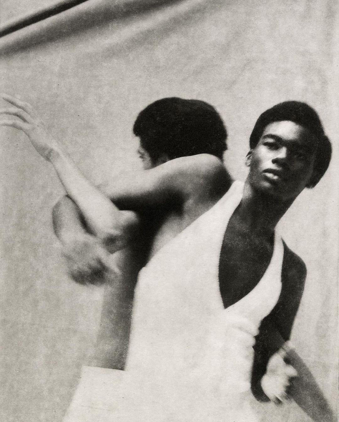 Striking black and white studio shot of a man in a white outfit with an arm raised, and an obscured reflection in the background.