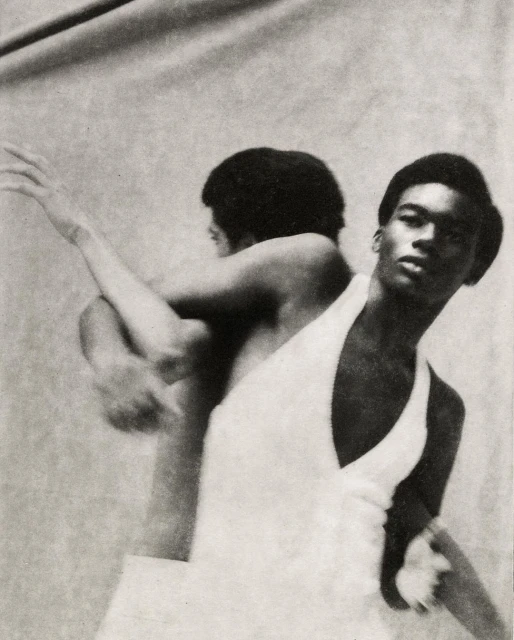 Striking black and white studio shot of a man in a white outfit with an arm raised, and an obscured reflection in the background.