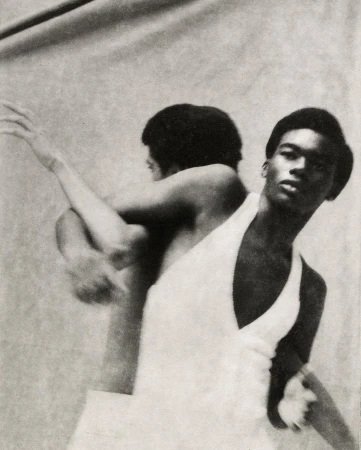 Striking black and white studio shot of a man in a white outfit with an arm raised, and an obscured reflection in the background.