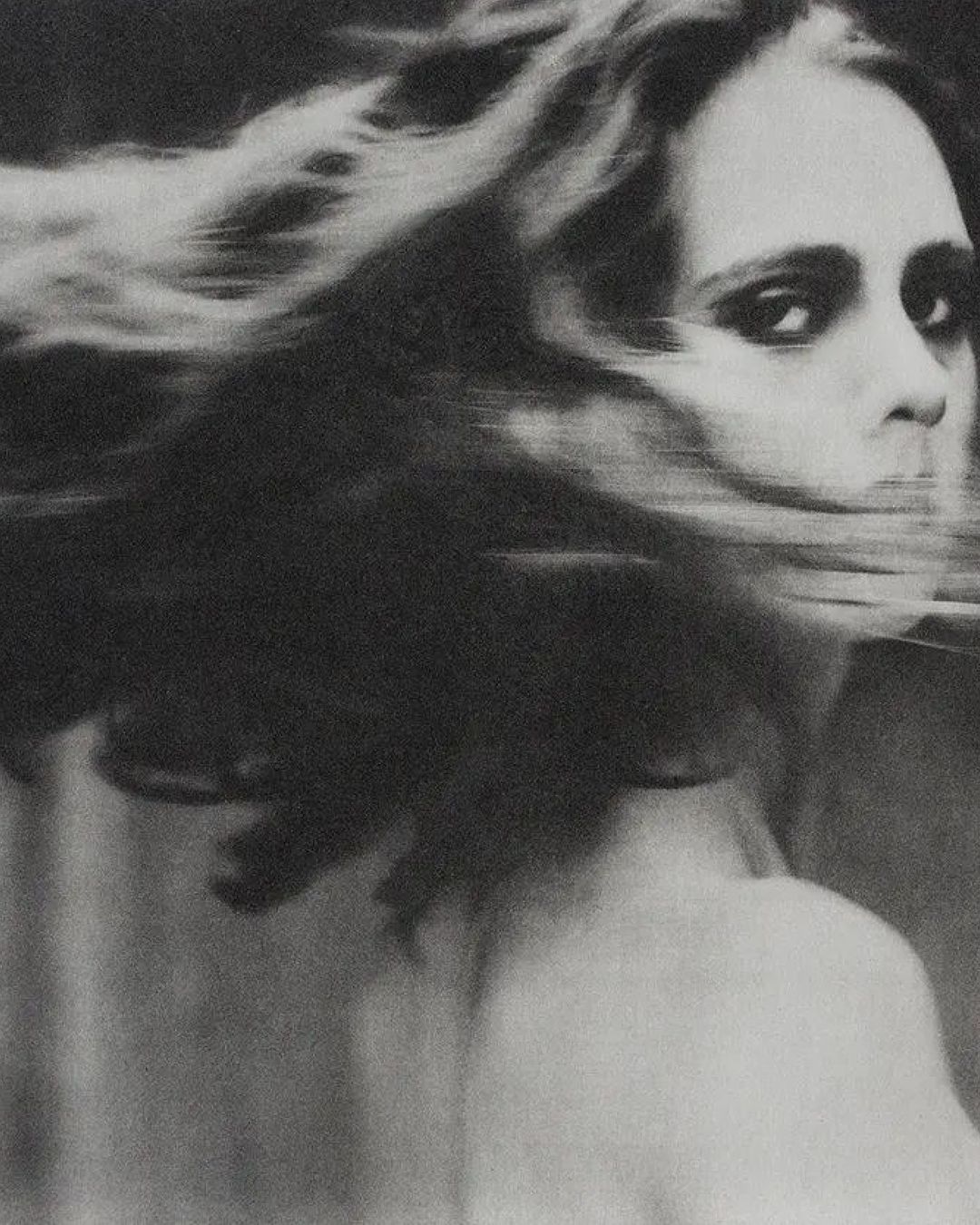 Black and white portrait of a woman with long, wind-blown hair, facing the camera with a neutral expression, wearing a dark choker necklace.