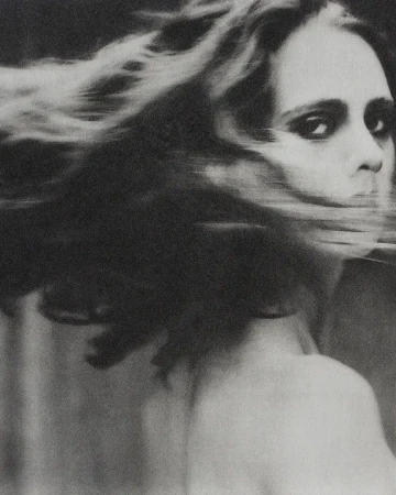 Black and white portrait of a woman with long, wind-blown hair, facing the camera with a neutral expression, wearing a dark choker necklace.
