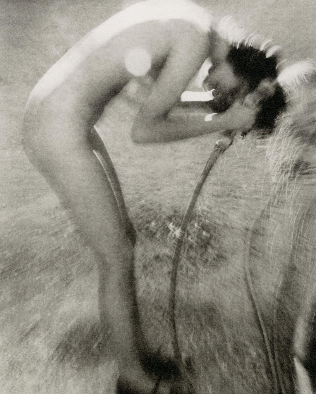Black and white image of an adult washing their face with a hose outdoors, featuring grassy ground in the background.