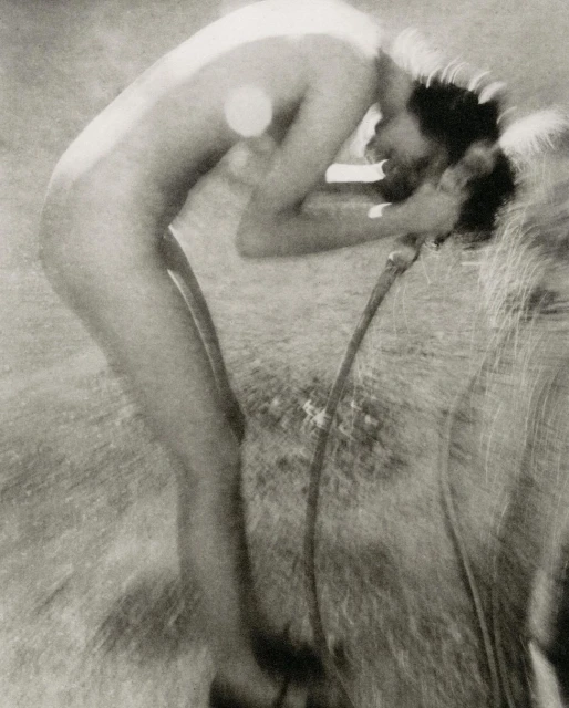 Black and white image of an adult washing their face with a hose outdoors, featuring grassy ground in the background.