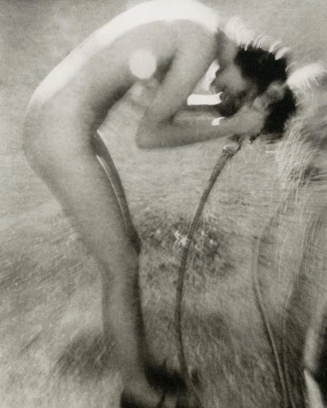 Black and white image of an adult washing their face with a hose outdoors, featuring grassy ground in the background.