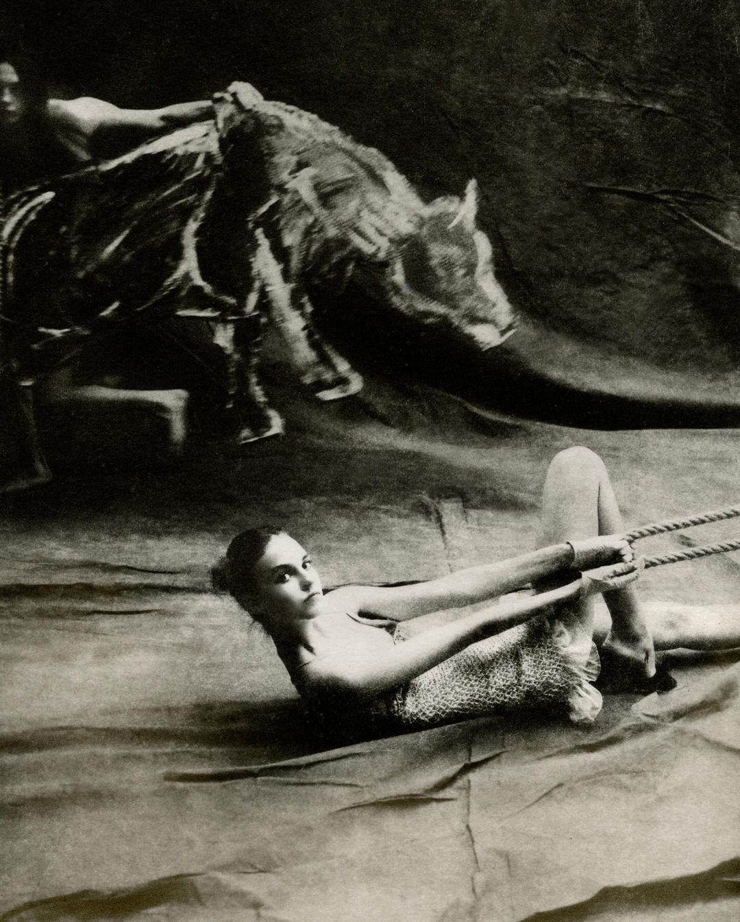 Striking black and white studio portrait of a woman lying down pulling rope, with a second person and animal overhead.