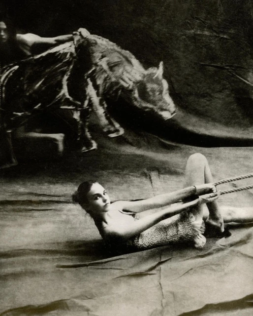 Striking black and white studio portrait of a woman lying down pulling rope, with a second person and animal overhead.