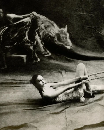 Striking black and white studio portrait of a woman lying down pulling rope, with a second person and animal overhead.