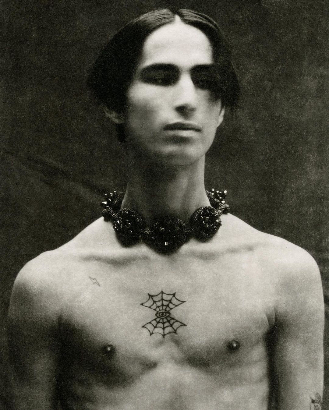 Monochrome portrait of an adult man with dark hair, a beaded necklace, and a spiderweb tattoo on his chest, set against a dark backdrop.