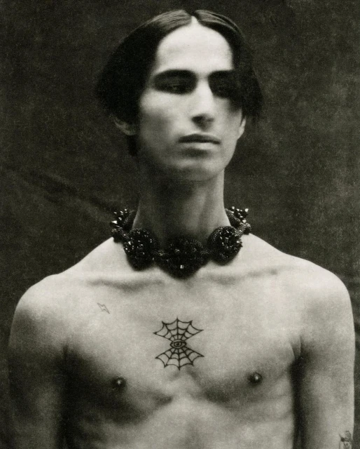 Monochrome portrait of an adult man with dark hair, a beaded necklace, and a spiderweb tattoo on his chest, set against a dark backdrop.