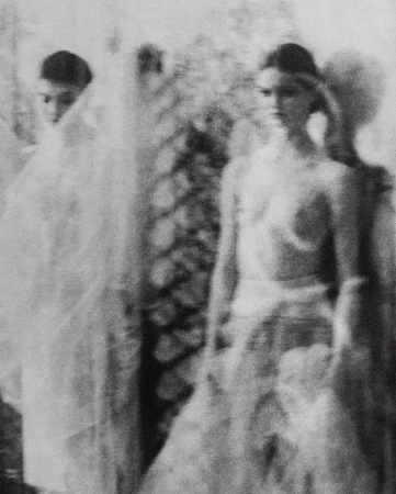 Two women in blurred black and white fashion portrait. One faces forward, the other is covered in white translucent material.