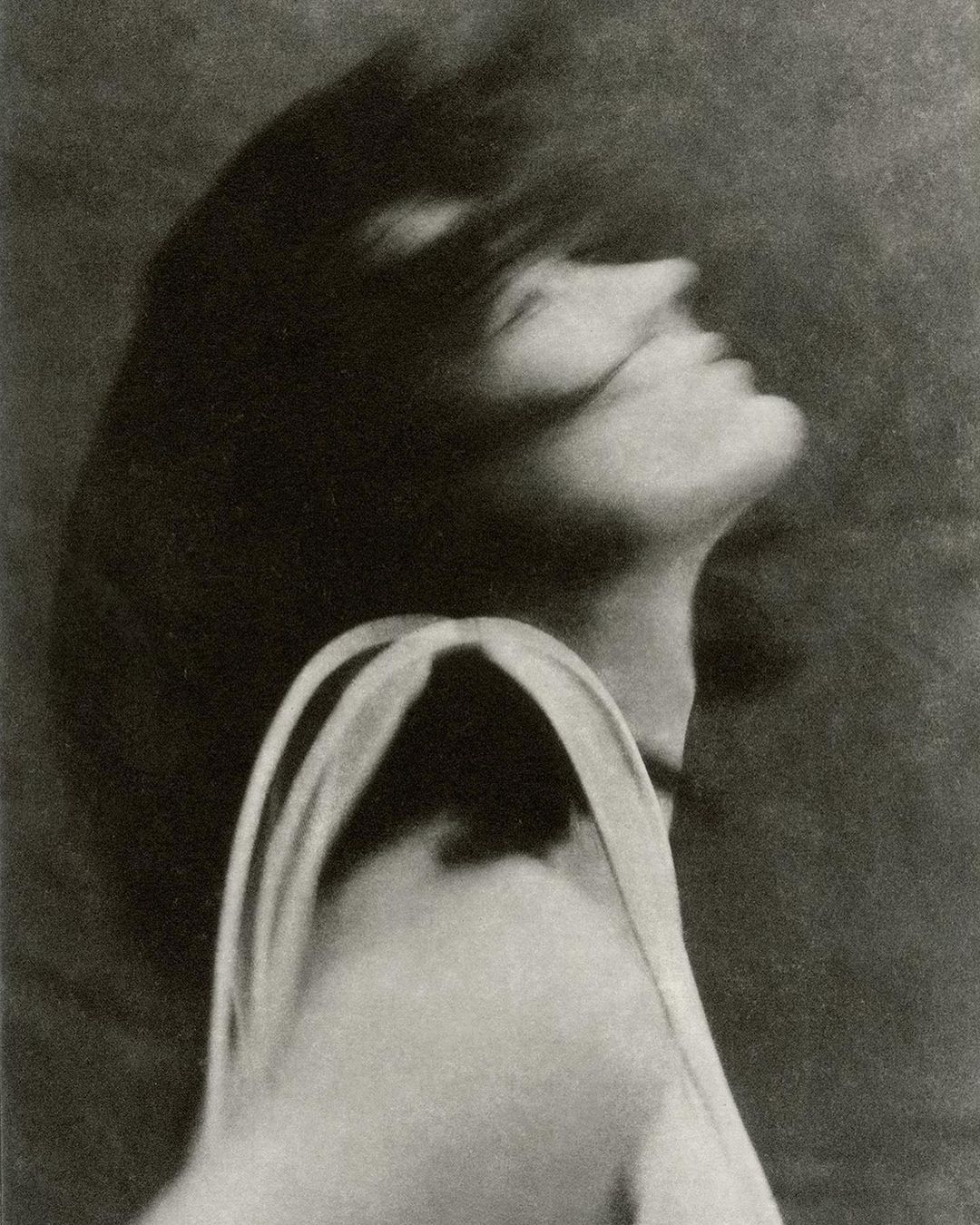 Monochrome portrait of a woman with flowing dark hair looking upwards, wearing a light-colored top against a neutral background.