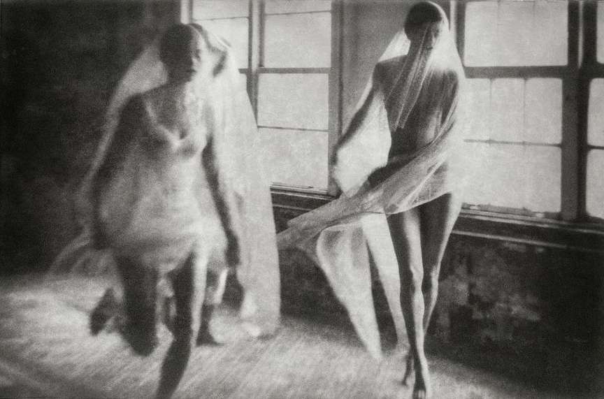 Black and white image of two women draped in sheer fabric, one in motion near a window, creating a sense of ethereal movement and mystery.