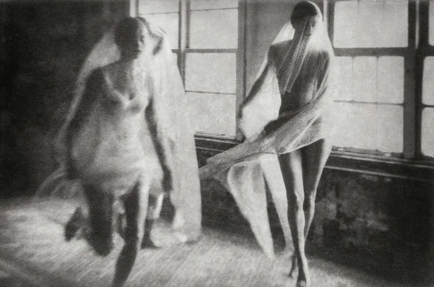 Black and white image of two women draped in sheer fabric, one in motion near a window, creating a sense of ethereal movement and mystery.