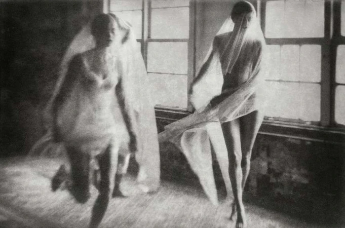 Black and white image of two women draped in sheer fabric, one in motion near a window, creating a sense of ethereal movement and mystery.