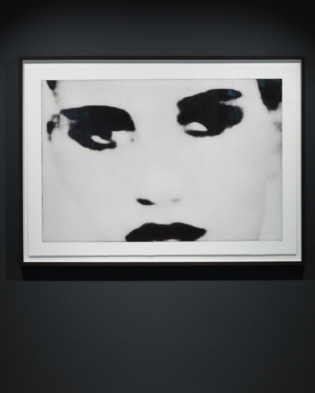Close-up black and white photo of a framed face in grayscale with dark eyes and lips, hanging on a dark grey wall, museum art exhibit.