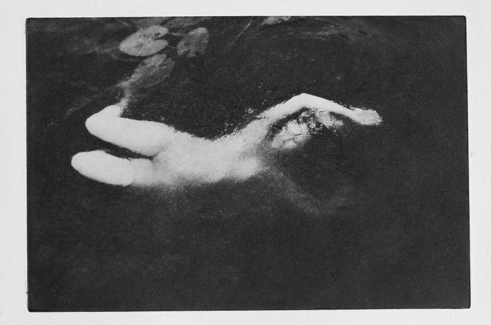 Monochrome photo of an adult female floating in dark water, lily pads in background; black-and-white ethereal photography.