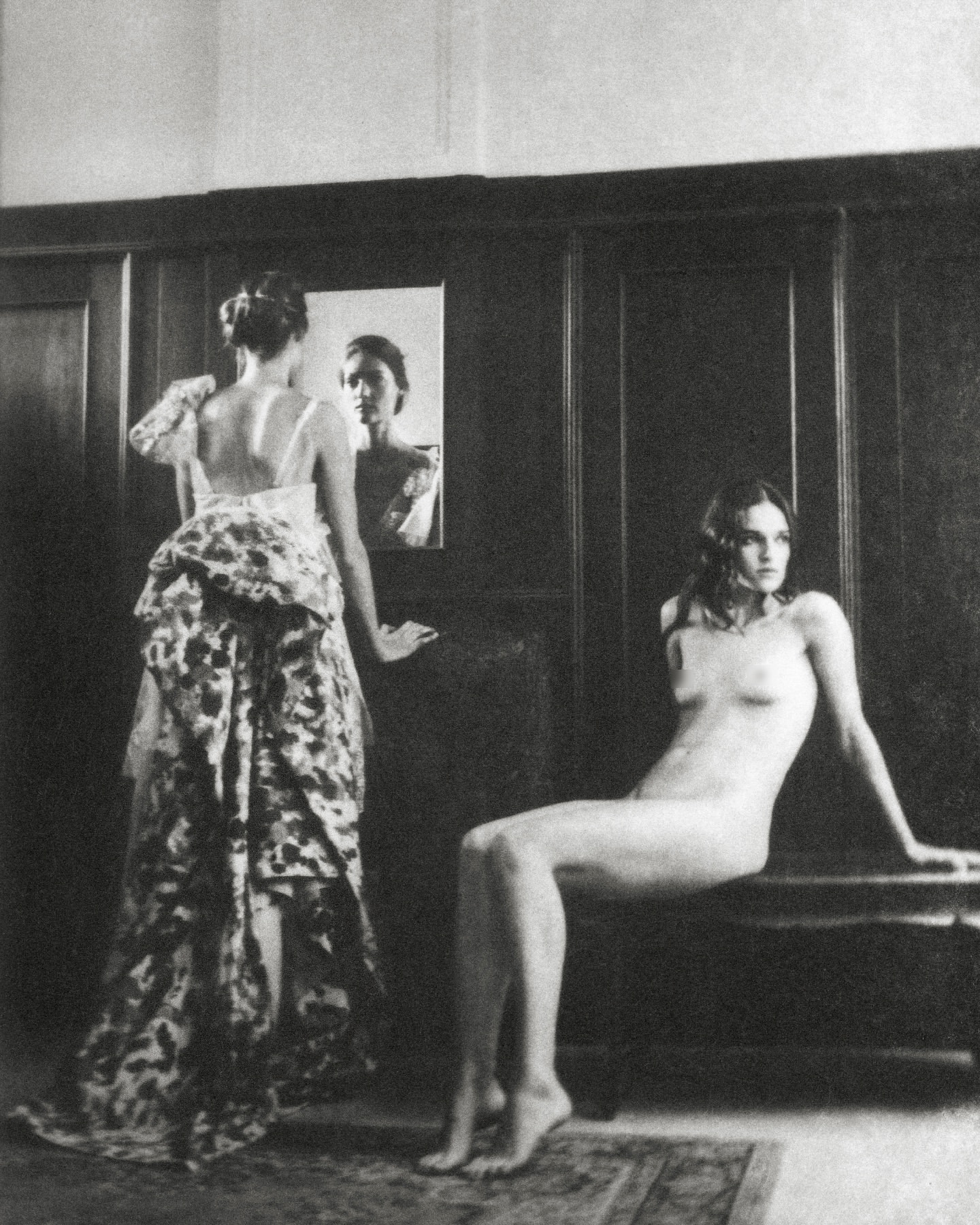 Monochrome photograph of two women indoors: one in a floral gown looking at a mirror and the other nude seated on a bench.