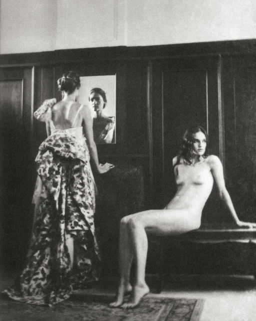 Monochrome photograph of two women indoors: one in a floral gown looking at a mirror and the other nude seated on a bench.